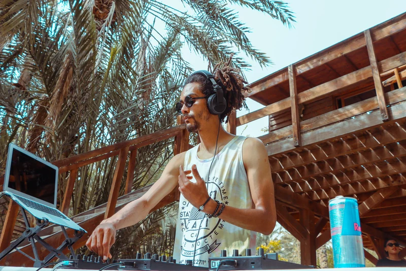 DJ JOK3R wearing headphones at an outdoor DJ setup beneath palm trees